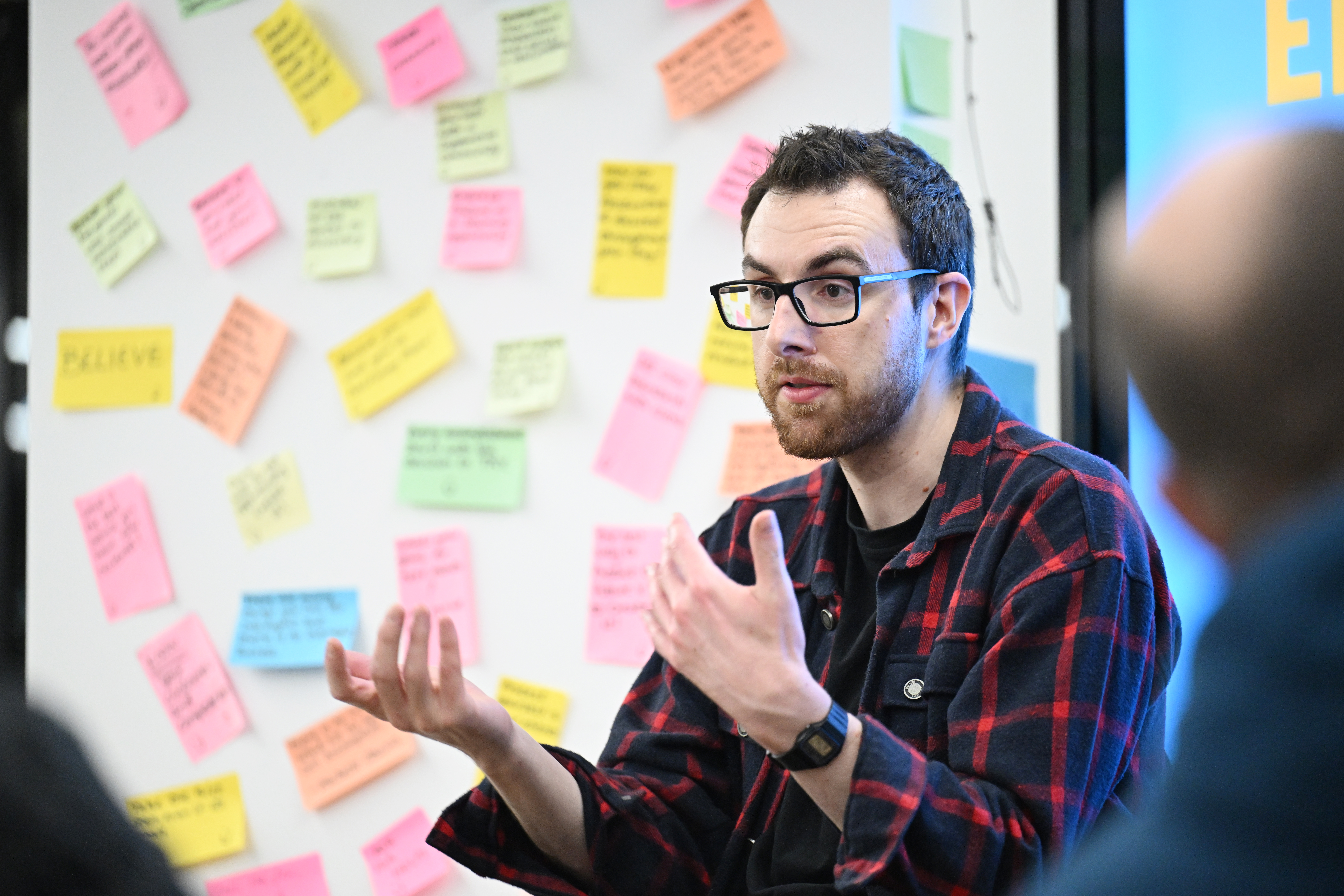 Person talking with lots of sticky notes on a wall behind them