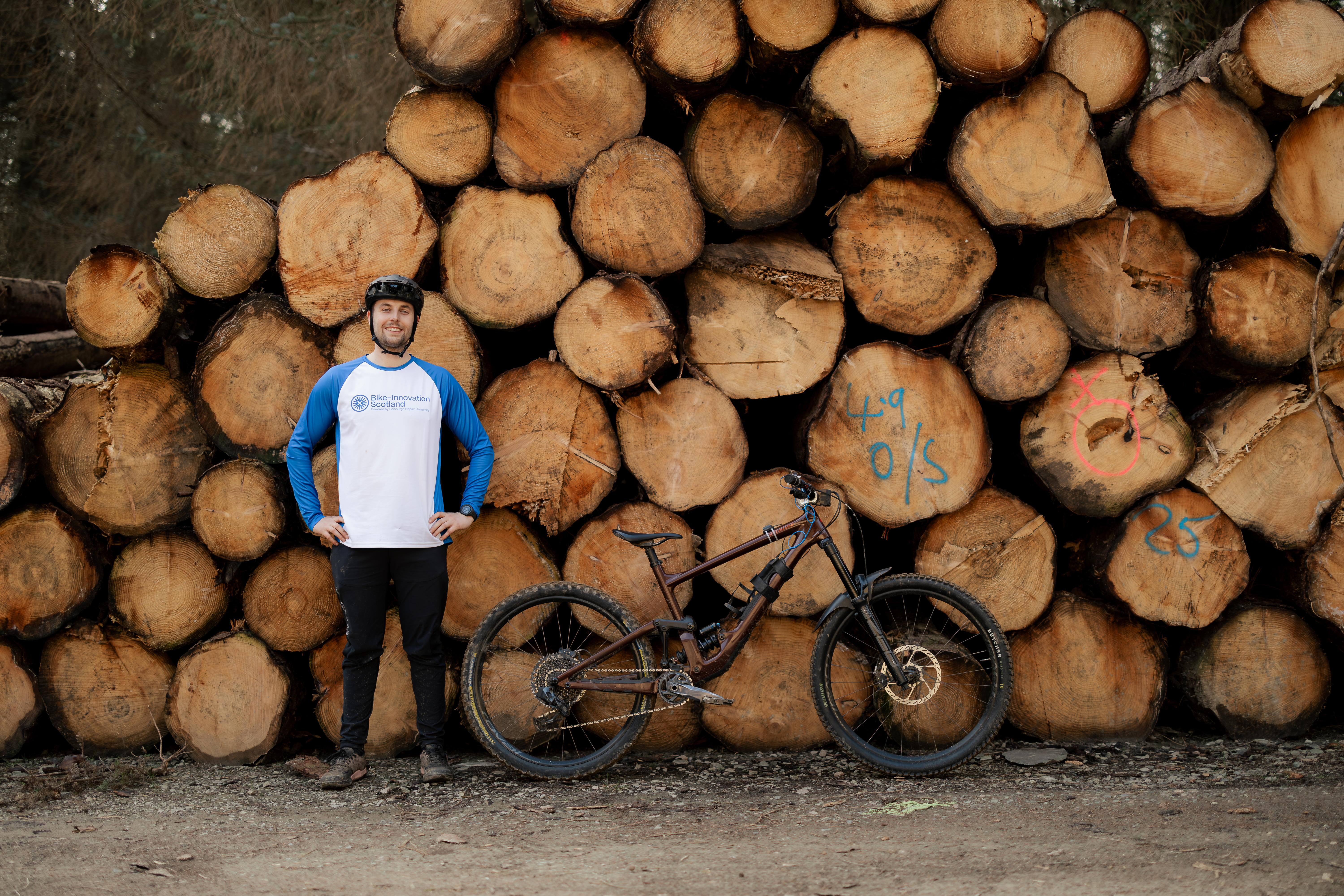 Stack of logs with a person smiling in front of it with their bike next them